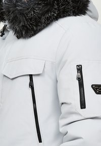 Veste d'hiver blanche avec capuche bordée de fourrure noire, fermetures éclair verticales noires sur les poches de la poitrine et des bras, et un petit logo de la marque sur la manche.