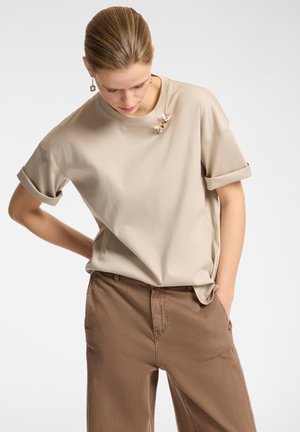 Beige T-Shirt mit kurzen Ärmeln und umgeschlagenen Säumen, verziert mit drei dekorativen Bienen-Anstecknadeln am Kragen. Kombiniert mit braunen Hosen.