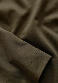 Olivgrüner Baumwollstoff mit einer glatten Textur, der sichtbare Nähte entlang der Kante und sanfte Falten aufweist.