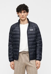 Geacă neagră matlasată, cu design matelassé, guler înalt și fermoar frontal. Logo EA7 pe pieptul stâng. Purtată peste un tricou alb.