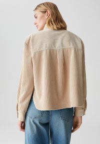 Maglione giacca in corduroy beige con una texture a coste, dotato di orlo arrotondato e polsini con bottoni, abbinato a jeans larghi blu chiaro.