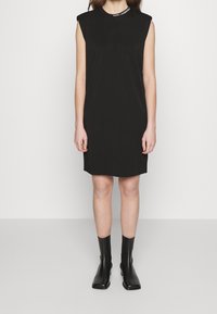 Czarna sukienka bez rękawów z okrągłym dekoltem, z subtelnym logo przy dekolcie. Materiał wydaje się gładki i dopasowany, do kolan.