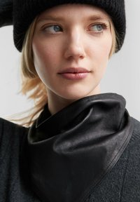 Schwarzer Leder-Schal, der um den Hals drapiert ist, mit einer glatten Textur und einem leichten Glanz, kombiniert mit einem dunklen Pullover und einer schwarzen Strickmütze.