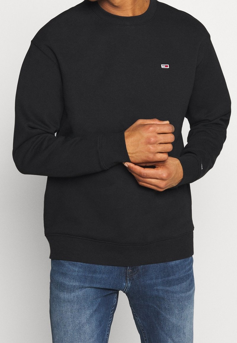 Sudadera de algodón negra con cuello redondo, puños y dobladillo acanalados, que presenta un pequeño parche con el logo en el pecho. Combinada con jeans azules.