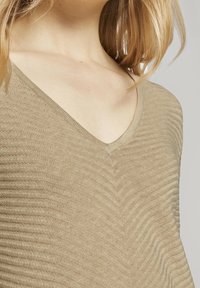 Beige V-Ausschnitt-Pullover mit gerippter Textur, der ein Zickzackmuster aufweist und aus weichem, leichtem Material gefertigt ist. Die enge Passform verbessert den Fall.