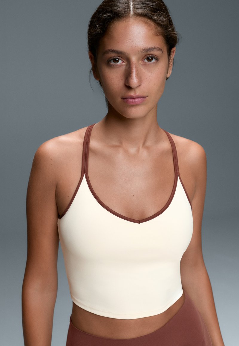 Young woman with brown hair tied back wearing a cream crop top with brown trim, standing against a plain gray background.