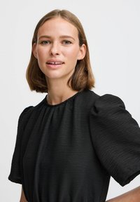 Čierne šaty s bufantovými rukávmi a nariaseným výstrihom. Tkanina má textúrovaný vzhľad. Modelka má vlasy strednej dĺžky a neutrálny výraz.