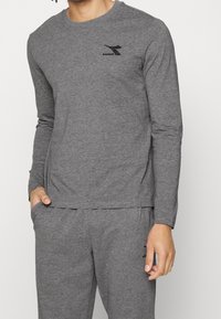 Harmaa pitkähihainen paita, joka on valmistettu pehmeästä puuvillasekoitekankaasta. Paidassa on pyöreä kaula-aukko ja pieni musta logo rinnassa.