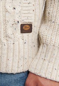 Maglione lavorato a maglia color crema con un motivo a trecce e coste, con un'etichetta in pelle marrone che riporta il logo "Petrol Industries."