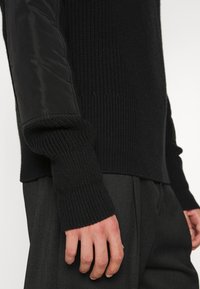 Personne portant un pull en maille noir avec des pièces en tissu matelassé sur les manches et un pantalon gris foncé, la main reposant le long du corps.