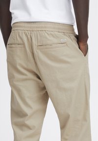 Pantaloni beige con vita elastica, dotati di due tasche posteriori, caratterizzati da una sottile texture e da un piccolo etichetta sulla tasca destra.