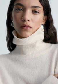 Maglione beige a collo alto con texture a coste e tessuto morbido, caratterizzato da un colletto alto. Orecchini a cerchio argentati completano il look.