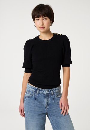 Zwarte gebreide trui met korte mouwen, ribbeltextuur en gouden knoopaccenten op de schouder, gecombineerd met lichtblauwe high-waisted jeans.