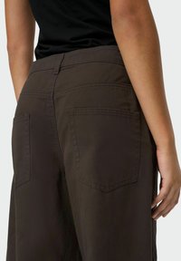 Pantalon en coton brun foncé avec une taille mi-haute, deux poches arrière et une texture douce. Coutures visibles et un design épuré et ajusté.