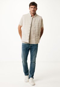 Light beige quilted vest with a stand collar, button closure, front pockets, worn over a beige polo shirt and blue jeans, completed with white sneakers.