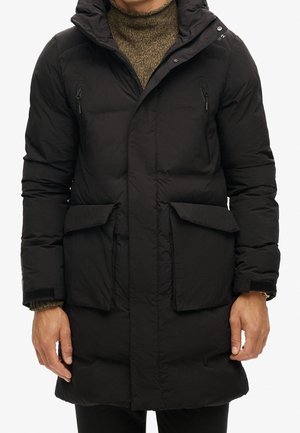 Homme portant un long manteau matelassé noir avec des poches poitrine zippées, de grandes poches à rabat et un pull à col roulé en maille marron en dessous.
