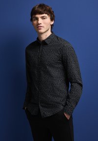 Černá košile s knoflíčky, pokrytá vzorem malých bílých a světle modrých čar, dlouhé rukávy a klasický límec, nošena s černými kalhotami.