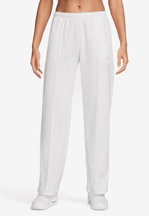 Persoon draagt witte sweatpants met een ontspannen pasvorm en een elastische tailleband en witte sneakers, staand tegen een effen achtergrond.