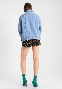 Chaqueta de mezclilla azul claro con un corte holgado, cuello clásico y dos bolsillos frontales, combinada con pantalones cortos negros con estampado floral y botas verdes hasta el tobillo.