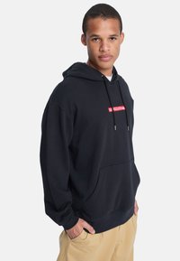 Sudadera negra de tejido suave, con un bolsillo frontal, capucha con cordón y un parche de logo rojo en el pecho. Diseño de ajuste casual.