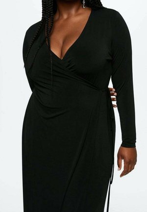 Kobieta o ciemnej karnacji i splecionych włosach, ubrana w czarną sukienkę kopertową z długim rękawem, głębokim dekoltem w serek i detalem z kokardą po bokach.