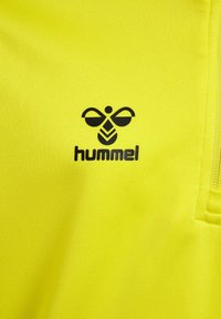 Heldere gele sportshirt gemaakt van gladde stof. Bevat een zwart logo met een gestileerd bijenmotief boven de tekst "hummel."