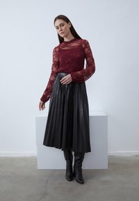 Burgundy csipkés hosszú ujjú felső egy fekete pliszírozott midi szoknyával párosítva. A modellen térdig érő fekete csizmák vannak. A háttér egyszínű világos árnyalatú.