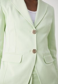 Blazer verde chiaro con un design strutturato, dotato di due bottoni con logo dorato, rever a punta e due tasche frontali. Tessuto liscio e su misura.