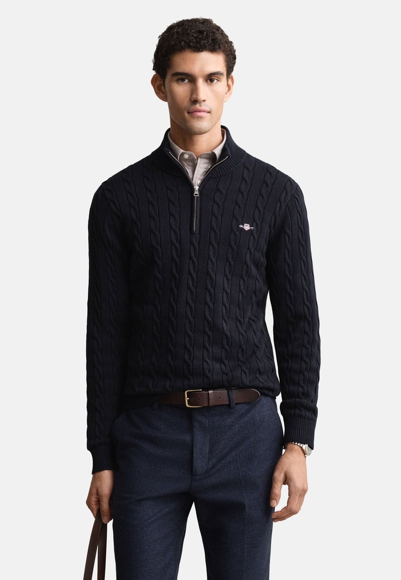 Námořnický svetr s copánkovým vzorem a čtvrtinovým zipem, s žebrovanými manžetami a lemem. Kombinován s tmavými kalhotami a hnědým koženým páskem.