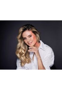 En kvinna med långt, vågigt blont hår bär en vit skjorta med knappar och ler. En röd linje går vertikalt längs hennes kind i bilden.