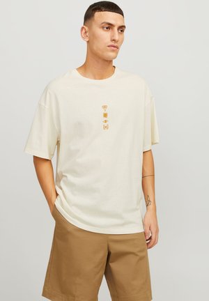 Lichtbeige katoenen T-shirt met oversized pasvorm, voorzien van kleine gouden grafische patronen: palm, zon, oog en vlinder gecentreerd op de borst.