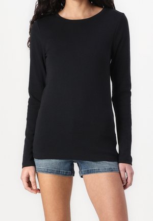 Frau trägt ein schlichtes schwarzes langärmliges Shirt und blaue Jeansshorts, steht vor weißem Hintergrund.