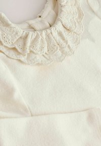 Indumento color crema con un colletto in pizzo arricciato decorato con ricami floreali e maniche lunghe realizzate in un tessuto morbido.