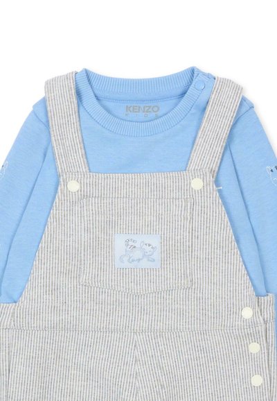 Camicia blu a maniche lunghe con scollo rotondo abbinata a salopette grigia a righe, caratterizzata da una tasca e dettagli con bottoni.