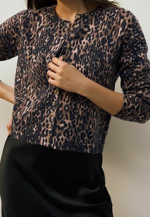 Femme portant un cardigan boutonné imprimé léopard et une jupe en satin noire, ajustant les boutons du cardigan avec la main.