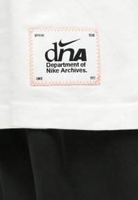 Λευκό ύφασμα πάνω σε ρούχο με μαύρο λογότυπο Nike και κείμενο «dna Department of Nike Archives» κεντημένο με πορτοκαλί κλωστή.