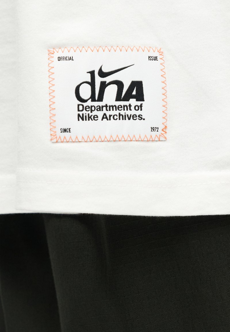 Weißes Stoffpatch auf Kleidung mit schwarzem Nike-Logo und dem Text "dna Department of Nike Archives", der mit orangefarbenem Faden aufgestickt ist.