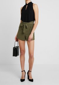 Top nero senza maniche con scollatura a chiave, abbinato a pantaloni corti verdi oliva a vita alta con cintura a fiocco, e tacchi a stiletto neri.