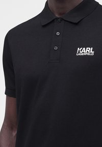 Gros plan sur un polo noir avec un col et trois boutons, présentant un logo texte blanc "KARL LAGERFELD" sur la poitrine gauche.