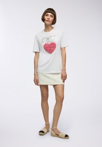 T-shirt bianco a maniche corte con un design a cuore texturizzato rosa, caratterizzato da scritte metalliche. Abbinato a una gonna color crema e espadrillas.
