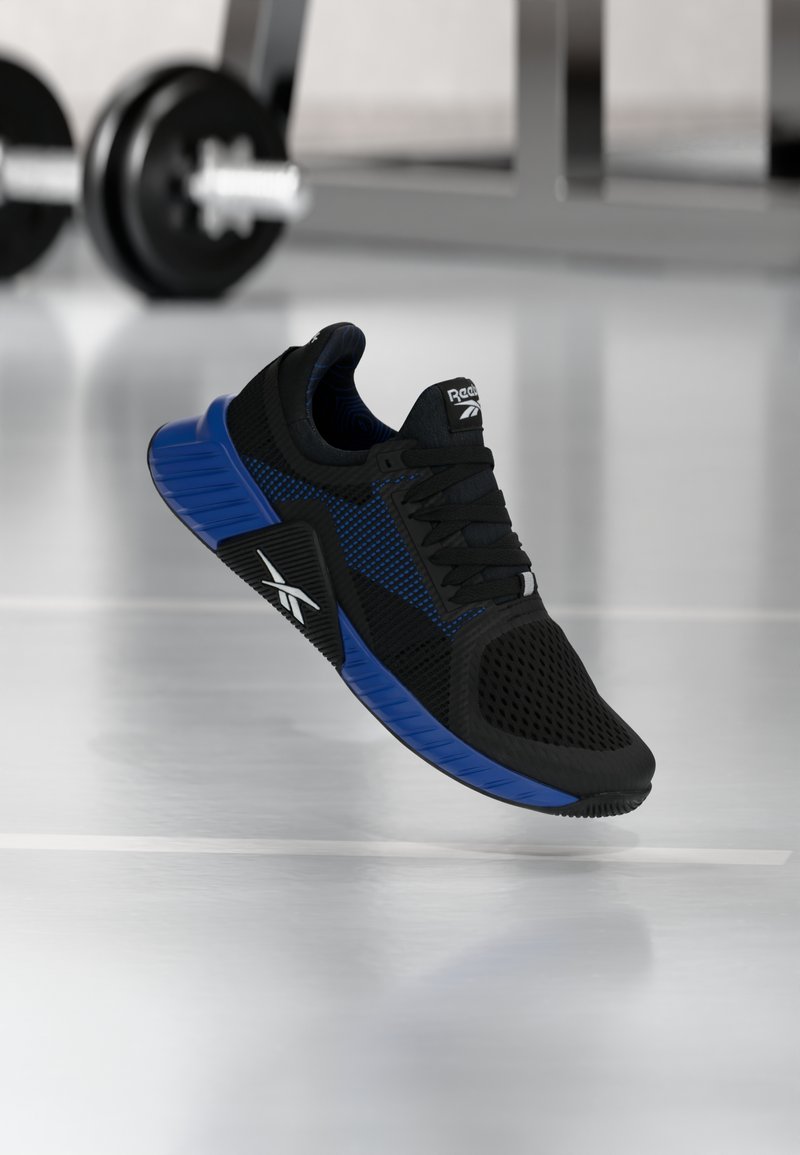 Μαύρο και μπλε αθλητικό παπούτσι Reebok κρεμασμένο πάνω από ανακλαστικό δάπεδο γυμναστηρίου, με θολά αλτήρες και εξοπλισμό γυμναστηρίου στο βάθος.