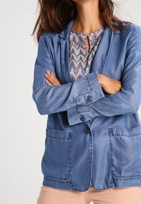 Niebieski blazer z denimu o luźnym kroju, z zapięciem na guziki i dwoma przednimi kieszeniami. Posiada klapowe wykończenie i gładką teksturę.