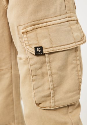 Light beige cargo pants with a side pocket, neat stitching, and a black label featuring the brand name "GARCA." Durable cotton material.