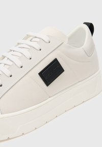 Baskets en toile blanches avec une épaisse semelle en caoutchouc blanc, accent avec étiquette noire et lacets plats blancs. Texture lisse et design minimaliste.