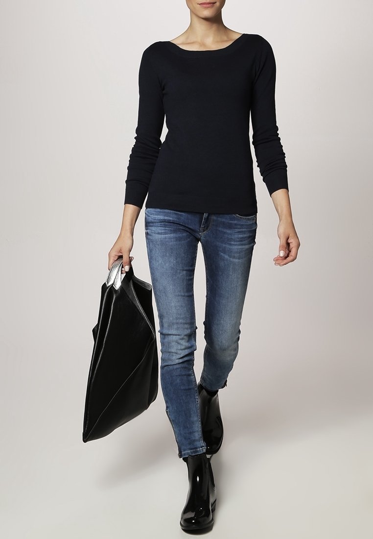 Schwarzes langärmeliges Oberteil, figurbetont, kombiniert mit blauen Jeans. Elegante schwarze Tasche in einer Hand gehalten, glänzende schwarze Stiefeletten.