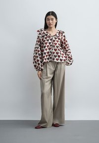 Vrouw staand, gekleed in beige wijde broek, bordeauxrode schoenen en een crème blouse met rode hartpatronen en opgeblazen lange mouwen.