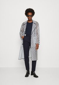 Transparenter, gemusterter langer Mantel mit marineblauen Akzenten, getragen über einem marineblauen Top und engen Hosen, kombiniert mit schwarzenHigh Heels und Sonnenbrille.