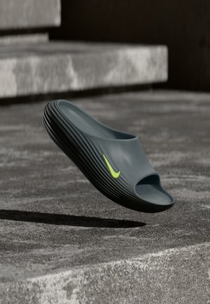 Jedna tmavosivá šľapka s textúrovanou čiernou podrážkou a žltým logom Nike, zachytená vo vzduchu nad betónovými schodmi.