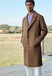 Brauner Wollmantel mit Reverskragen, vorderem Knopfverschluss und Seitentaschen, getragen über einem lila Pullover und kombiniert mit weißen Hosen.