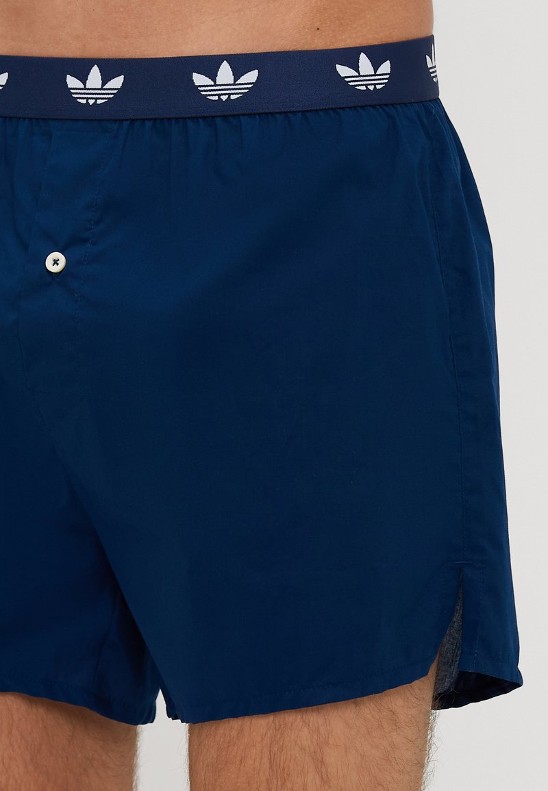 Mørkeblå badebukser lavet af letvægtsmateriale, med en elastisk talje og hvide logo-detaljer samt en knaplukning foran.